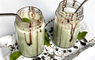 two chocolate mint shakes with straws in mason jars on a decorative tray dribbled in chocolate with mint leaves. white background.