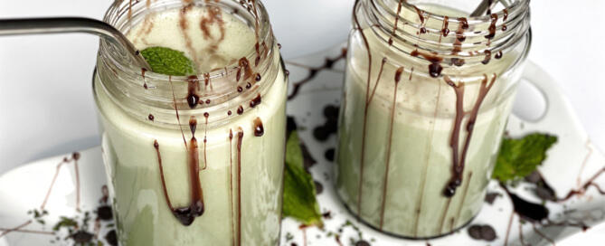 two chocolate mint shakes with straws in mason jars on a decorative tray dribbled in chocolate with mint leaves. white background.